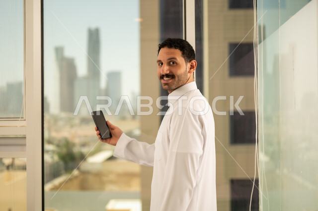 استخدام جهاز تقني حديث ومتطور،  صورة مقربة من الخلف لرجل عربي خليجي سعودي يرتدي الثوب التقليدي يعمل على الهاتف المحمول، التقدم والتطور المعماري في دول الخليج، خلفية الأبراج وناطحات السحاب في مدينة الرياض
