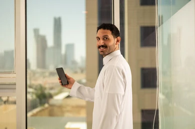 استخدام جهاز تقني حديث ومتطور،  صورة مقربة من الخلف لرجل عربي خليجي سعودي يرتدي الثوب التقليدي يعمل على الهاتف المحمول، التقدم والتطور المعماري في دول الخليج، خلفية الأبراج وناطحات السحاب في مدينة الرياض