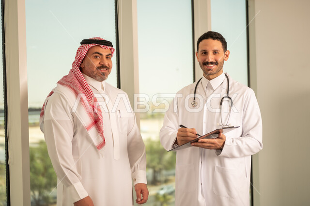  تدوين نتائج الفحوصات بالسجل الطبي لمريض كبير بالسن، حسن التعامل مع المراجعين، ايماءات البشاشة والسرور، طبيب عربي خليجي سعودي يرتدي الثوب التقليدي والمعطف الابيض يحمل بيده ملفات المرضى، العمل في القطاع الصحي, اعداد التقارير الطبية