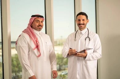  تدوين نتائج الفحوصات بالسجل الطبي لمريض كبير بالسن، حسن التعامل مع المراجعين، ايماءات البشاشة والسرور، طبيب عربي خليجي سعودي يرتدي الثوب التقليدي والمعطف الابيض يحمل بيده ملفات المرضى، العمل في القطاع الصحي, اعداد التقارير الطبية