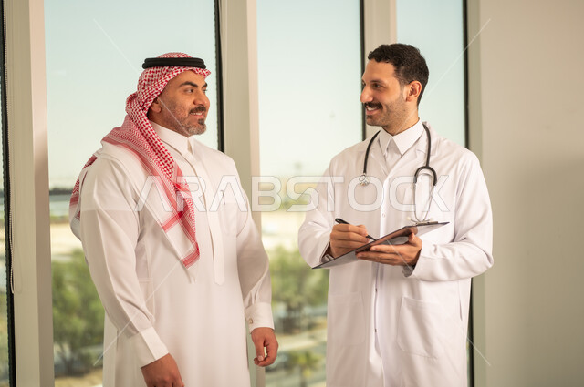 اعداد التقارير الطبية، تدوين نتائج الفحوصات بالسجل الطبي لمريض كبير بالسن، حسن التعامل مع المراجعين، ايماءات البشاشة والسرور، طبيب عربي خليجي سعودي يرتدي الثوب التقليدي والمعطف الابيض يحمل بيده ملفات المرضى، العمل في القطاع الصحي