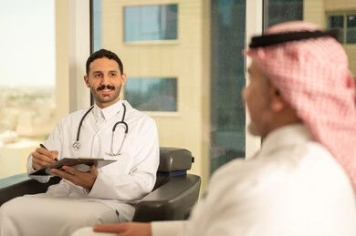 اعداد التقارير الطبية، تدوين نتائج الفحوصات بالسجل الطبي لمريض كبير بالسن، حسن التعامل مع المراجعين، ايماءات البشاشة والسرور، طبيب عربي خليجي سعودي يرتدي الثوب التقليدي والمعطف الابيض يحمل بيده ملفات المرضى، العمل في القطاع الصحي