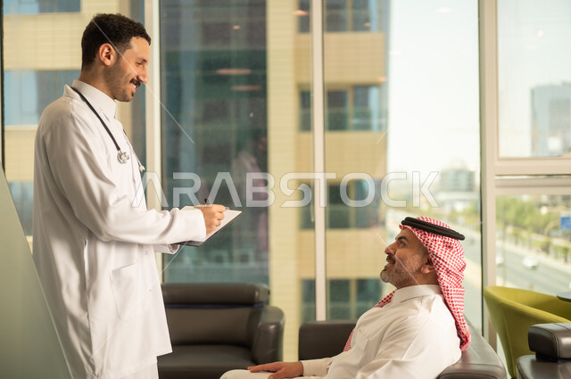 اعداد التقارير الطبية، تدوين نتائج الفحوصات بالسجل الطبي لمريض كبير بالسن، حسن التعامل مع المراجعين، ايماءات البشاشة والسرور، طبيب عربي خليجي سعودي يرتدي الثوب التقليدي والمعطف الابيض يحمل بيده ملفات المرضى، العمل في القطاع الصحي