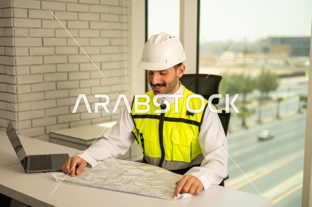 Working in the engineering field, the concept of engineering and construction, a Saudi Gulf Arab engineer wearing a safety vest and helmet sitting at the desk working on the engineering plan, reviewing and auditing engineering plans, studying the fundamentals of the project, completing the drawing of construction blueprints, using modern technical devices.