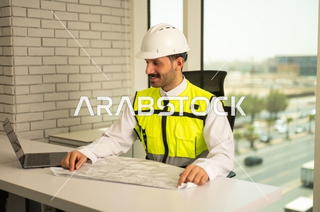 Study of project fundamentals, auditing and reviewing engineering plans, the concept of engineering and construction, a Saudi Gulf Arab engineer wearing a vest and safety helmet sitting at a desk working on the engineering plan, completing the drawing of construction blueprints, using modern technical devices, youth professions and jobs.
