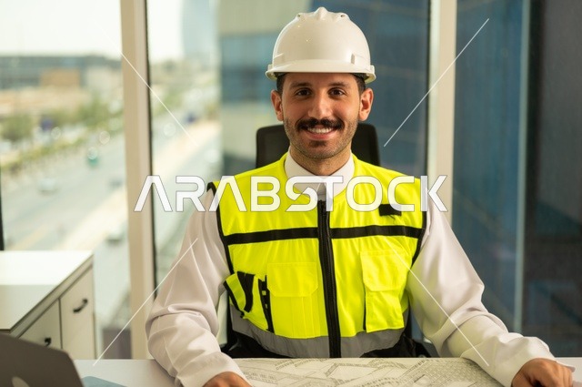 تدقيق ومراجعة الخطط الهندسية، مفهوم الهندسة والإنشاءات، صورة مقربة لمهندس عربي خليجي سعودي يرتدي سترة وخوذة الحماية يجلس على المكتب يعمل على المخطط الهندسي، إكمال رسم مخطوطات البناء، استخدام الأجهزة التقنية الحديثة، مهن ووظائف شبابية