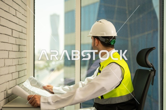 الإعمار بسواعد أبناء الوطن،  مفهوم الهندسة والإنشاءات، صورة جانبية  لمهندس عربي خليجي سعودي يرتدي سترة وخوذة الحماية يجلس على المكتب يتأمل التطور العمراني للمباني، تدقيق ومراجعة الخطط الهندسية، دراسة أساسيات المشروع، إكمال رسم مخطوطات البناء