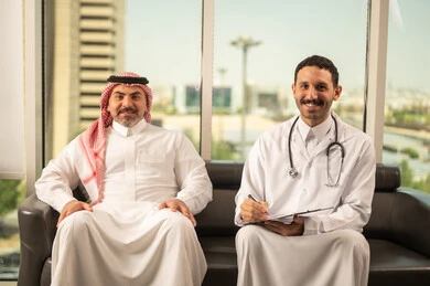 اعداد التقارير الطبية، تدوين نتائج الفحوصات بالسجل الطبي لمريض كبير بالسن، حسن التعامل مع المراجعين، ايماءات البشاشة والسرور، طبيب عربي خليجي سعودي يرتدي الثوب التقليدي والمعطف الابيض يحمل بيده ملفات المرضى، العمل في القطاع الصحي