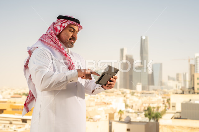 جهاز تقني حديث ومتطور، رجل عربي خليجي سعودي يرتدي الثوب والشماغ التقليدي يقوم باستخدام جهاز التابلت اللوحي، إنجاز العمل عن بعد، خلفية الأبراج وناطحات السحاب في مدينة الرياض, تصفح مواقع التواصل الاجتماعي
