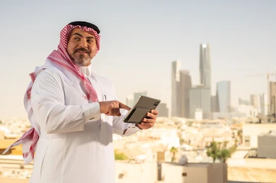 جهاز تقني حديث ومتطور، رجل عربي خليجي سعودي يرتدي الثوب والشماغ التقليدي يقوم باستخدام جهاز التابلت اللوحي، إنجاز العمل عن بعد، خلفية الأبراج وناطحات السحاب في مدينة الرياض, تصفح مواقع التواصل الاجتماعي