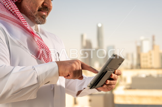 جهاز تقني حديث ومتطور، رجل عربي خليجي سعودي يرتدي الثوب والشماغ التقليدي يقوم باستخدام جهاز التابلت اللوحي، إنجاز العمل عن بعد، خلفية الأبراج وناطحات السحاب في مدينة الرياض, تصفح مواقع التواصل الاجتماعي