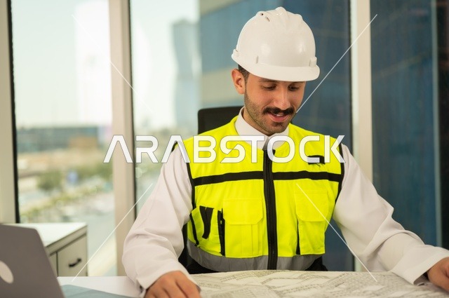 تدقيق ومراجعة الخطط الهندسية، مفهوم الهندسة والإنشاءات، صورة مقربة لمهندس عربي خليجي سعودي يرتدي سترة وخوذة الحماية يجلس على المكتب يعمل على المخطط الهندسي، إكمال رسم مخطوطات البناء، استخدام الأجهزة التقنية الحديثة، مهن ووظائف شبابية