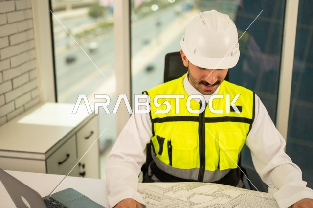 Youth professions and jobs, auditing and reviewing engineering plans, studying project fundamentals, the concept of engineering and construction, a Saudi Gulf Arab engineer wearing a vest and safety helmet sitting at the desk working on the engineering plan, completing the drawing of construction blueprints, using modern technical devices.