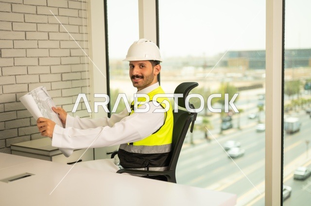 تدقيق ومراجعة الخطط الهندسية، الإعمار بسواعد أبناء الوطن،  مفهوم الهندسة والإنشاءات، صورة جانبية  لمهندس عربي خليجي سعودي مبتسم يرتدي سترة وخوذة الحماية يجلس على المكتب يحمل المخططات الهندسية، دراسة أساسيات المشروع، إكمال رسم مخطوطات البناء