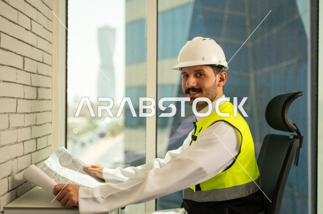 صورة جانبية  لمهندس عربي خليجي سعودي مبتسم يرتدي سترة وخوذة الحماية يجلس على المكتب يحمل المخططات الهندسية،  تدقيق ومراجعة الخطط الهندسية، الإعمار بسواعد أبناء الوطن،  مفهوم الهندسة والإنشاءات، دراسة أساسيات المشروع، إكمال رسم مخطوطات البناء