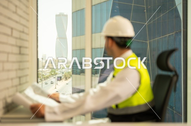 الإعمار بسواعد أبناء الوطن،  مفهوم الهندسة والإنشاءات، صورة جانبية  لمهندس عربي خليجي سعودي يرتدي سترة وخوذة الحماية يجلس على المكتب يتأمل التطور العمراني للمباني، تدقيق ومراجعة الخطط الهندسية، دراسة أساسيات المشروع، إكمال رسم مخطوطات البناء