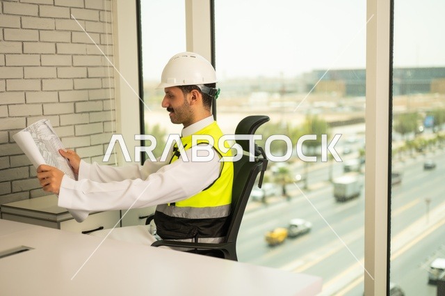 متابعة مخطط الرسوم الهندسية، الإعمار بسواعد أبناء الوطن،  مفهوم الهندسة والإنشاءات، صورة جانبية  لمهندس عربي خليجي سعودي يرتدي سترة وخوذة الحماية يجلس على المكتب يحمل المخططات الهندسية، دراسة أساسيات المشروع، إكمال رسم مخطوطات البناء