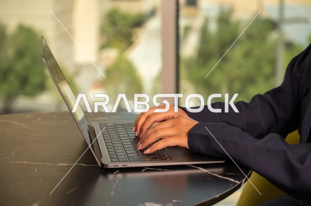 ادارة الأعمال عن بعد، استخدام جهاز تقني حديث ومتطور، صورة مقربة لامرأة عربية خليجية سعودية ترتدي العباءة والحجاب تقوم باستخدام الحاسوب المحمول في المكتب، دمج التكنولوجيا بالعمل، إنجاز المهام المطلوبة عبر اللابتوب، مهنة ووظيفة نسائية مكتبية