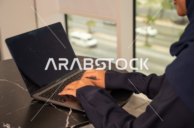 Remote business management, using a modern and advanced technical device, a close-up image of a Saudi Gulf Arab woman wearing an abaya and hijab using a laptop in the office, integrating technology with work, completing required tasks via the laptop, a female office profession and job.