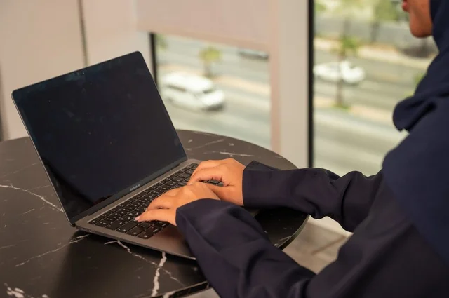 ادارة الأعمال عن بعد، استخدام جهاز تقني حديث ومتطور، صورة مقربة لامرأة عربية خليجية سعودية ترتدي العباءة والحجاب تقوم باستخدام الحاسوب المحمول في المكتب، دمج التكنولوجيا بالعمل، إنجاز المهام المطلوبة عبر اللابتوب، مهنة ووظيفة نسائية مكتبية