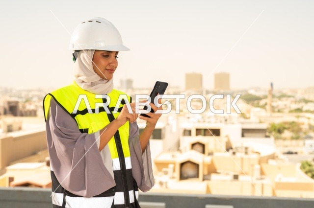 العمل في المجال الهندسي, مهندسة عربية خليجية سعودية مبتسمة محجبة ترتدي الخوذة وسترة الحماية تقف باستقامة وتعمل على الهاتف المحمول ، مفهوم الهندسة و الإنشاءات, مهن ووظائف نسائية هندسية, خلفية أبراج وناطحات سحاب في الرياض