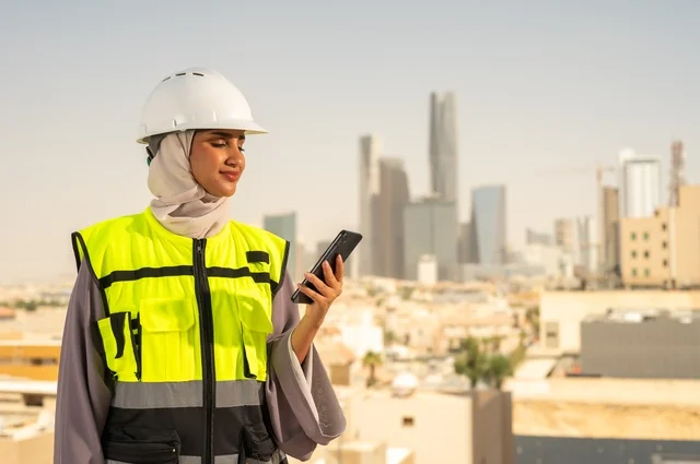 Working in the engineering field, a smiling, veiled Arab Gulf Saudi engineer wearing a helmet and safety vest stands upright and works on a mobile phone, the concept of engineering and construction, engineering jobs and professions for women, with a background of towers and skyscrapers in Riyadh.