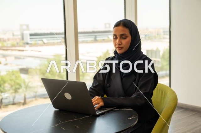 Integrating technology into work, remote business management, using a modern and advanced technical device, an Arab Gulf Saudi woman wearing an abaya and hijab using a laptop in the office, completing the required tasks via the laptop, a female office profession and job.