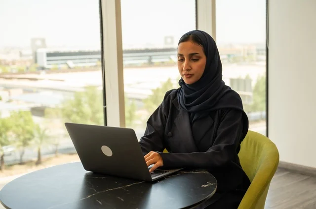 Integrating technology into work, remote business management, using a modern and advanced technical device, an Arab Gulf Saudi woman wearing an abaya and hijab using a laptop in the office, completing the required tasks via the laptop, a female office profession and job.