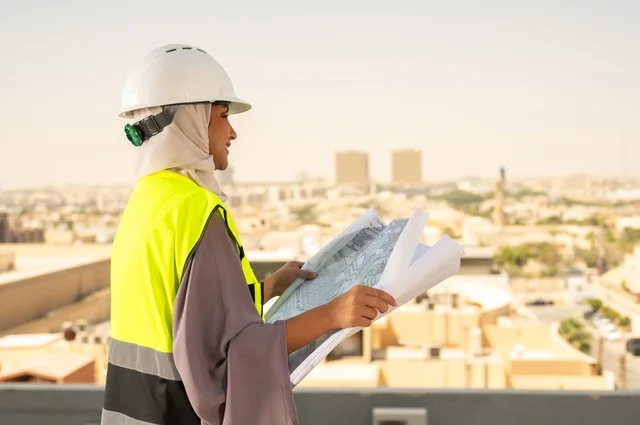 مهندسة معمارية عربية سعودية خليجية تحمل بيدها مخطط البناء، مهندسة انشاءات محجبة ترتدي بدلة السلامة في العمل ، خوذة العمل، لباس رسمي للعمل، مشروع بناء، مهن ووظائف نسائية هندسية, ايماءات وجه تدل على السرور، العمل في المجال الهندسي, مفهوم الهندسة و الإنشاءات