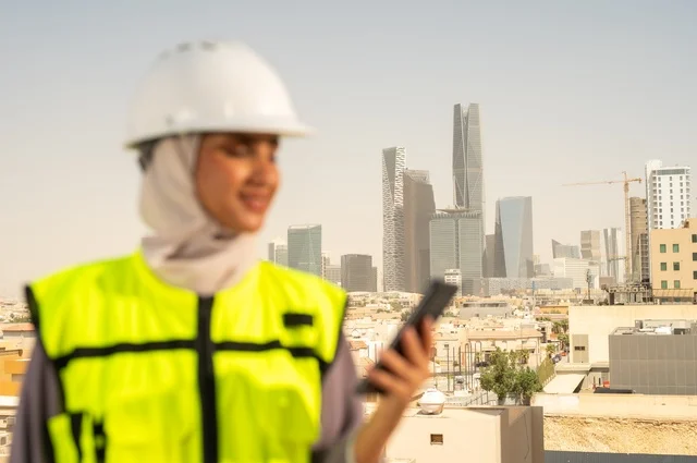 ايماءات وجه تدل على السرور، مفهوم الهندسة و الإنشاءات, صورة مقربة لمهندسة عربية خليجية سعودية مبتسمة محجبة ترتدي الخوذة وسترة الحماية تقف باستقامة تحمل على هاتف محمول ، العمل في المجال الهندسي, مهن ووظائف نسائية هندسية, خلفية أبراج وناطحات سحاب في الرياض