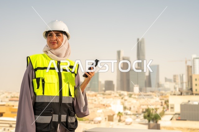 مفهوم الهندسة و الإنشاءات, مهندسة عربية خليجية سعودية مبتسمة محجبة ترتدي الخوذة وسترة الحماية تقف باستقامة وتنظر إلى الكاميرا بثقة تحمل على هاتف محمول ، العمل في المجال الهندسي, مهن ووظائف نسائية هندسية, خلفية أبراج وناطحات سحاب في الرياض