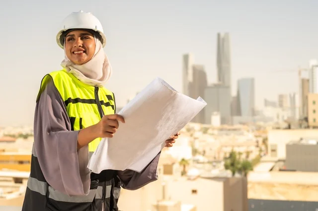 Gestures of joy and pride, an Arab Saudi Gulf architectural engineer holding a building plan in her hand, a hijab-wearing construction engineer dressed in a work safety suit, a work helmet, formal work attire, a construction project, engineering professions and jobs for women, facial expressions indicating joy, the concept of engineering and construction.