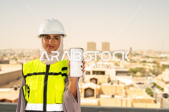Engineering jobs and professions for women, facial expressions indicating happiness, a smiling Arab Gulf Saudi engineer wearing a hijab, helmet, and safety vest standing upright displaying a white mobile phone background, working in the engineering field, the concept of engineering and construction, background of towers and skyscrapers in Riyadh.