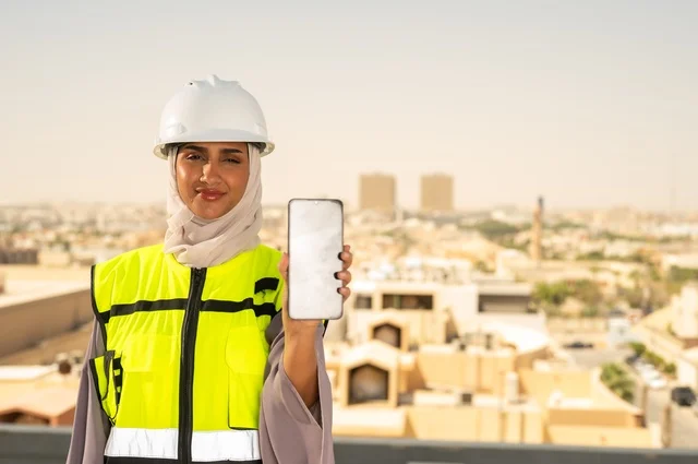 Engineering jobs and professions for women, facial expressions indicating happiness, a smiling Arab Gulf Saudi engineer wearing a hijab, helmet, and safety vest standing upright displaying a white mobile phone background, working in the engineering field, the concept of engineering and construction, background of towers and skyscrapers in Riyadh.