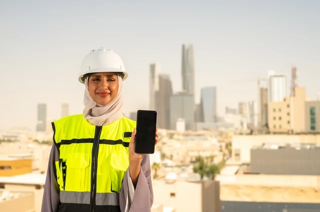 Background of towers and skyscrapers in Riyadh, facial expressions indicating joy, the concept of engineering and construction, a smiling, veiled Saudi Gulf Arab engineer wearing a helmet and safety vest standing upright displaying a black mobile phone background, working in the engineering field, engineering professions and jobs for women.