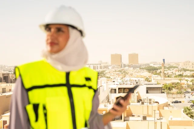 Facial expressions indicating joy, the concept of engineering and construction, a smiling, veiled Arab Gulf Saudi engineer wearing a helmet and safety vest standing upright holding a mobile phone, working in the engineering field, engineering jobs and professions for women, background of towers and skyscrapers in Riyadh.