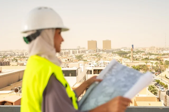 صورة جانبية مقربة لمهندسة معمارية عربية سعودية خليجية تحمل بيدها مخطط البناء، مهندسة انشاءات محجبة ترتدي بدلة السلامة في العمل ، خوذة العمل، لباس رسمي للعمل، مهن ووظائف نسائية هندسية, ايماءات وجه تدل على السرور، مفهوم الهندسة و الإنشاءات