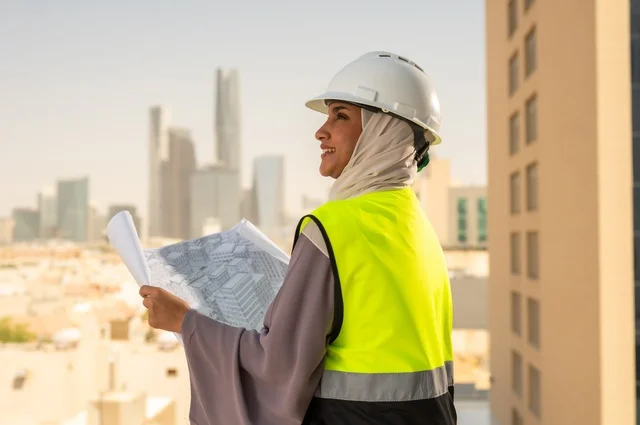 مفهوم الهندسة و الإنشاءات، صورة جانبية لمهندسة معمارية عربية سعودية خليجية تحمل بيدها مخطط البناء، مهندسة انشاءات محجبة ترتدي بدلة السلامة في العمل ، خوذة العمل، لباس رسمي للعمل، مهن ووظائف نسائية هندسية, ايماءات وجه تدل على السرور،