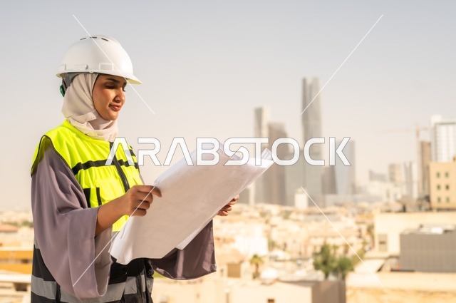 مشروع بناء، صورة جانبية لمهندسة معمارية عربية سعودية خليجية تحمل بيدها مخطط البناء، مهندسة انشاءات محجبة ترتدي بدلة السلامة في العمل ، خوذة العمل، لباس رسمي للعمل، مهن ووظائف نسائية هندسية، خلفية أبراج وناطحات سحاب