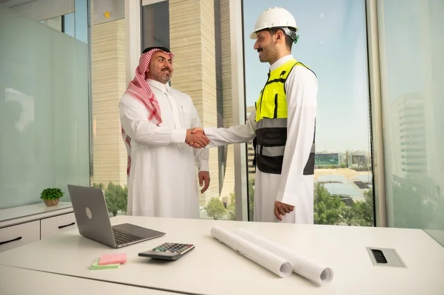 The handshake, greeting, and reception, the concept of cooperation and joint work, a Saudi Gulf Arab engineer wearing a helmet and safety vest shaking hands with a businessman wearing a ghutrah and traditional thobe, agreeing on engineering plans and projects at the work site, architectural constructions in Saudi lands.