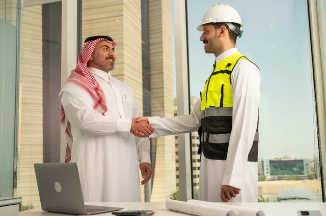 المصافحة والترحيب والاستقبال, مفهوم التعاون والعمل المشترك, مهندس عربي خليجي سعودي يرتدي الخوذة وسترة الحماية يقوم بمصافحة رجل اعمال يرتدي الشماغ و الثوب التقليدي، الاتفاق على الخطط والمشاريع الهندسية في موقع العمل , الإنشاءات المعمارية في الأراضي السعودية