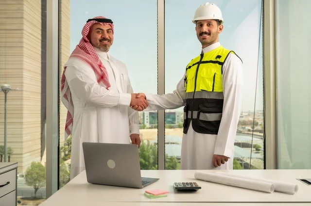 The handshake, greeting, and reception, the concept of cooperation and joint work, a Saudi Gulf Arab engineer wearing a helmet and safety vest shaking hands with a businessman wearing a ghutrah and traditional thobe, agreeing on engineering plans and projects at the work site, architectural constructions in Saudi lands.