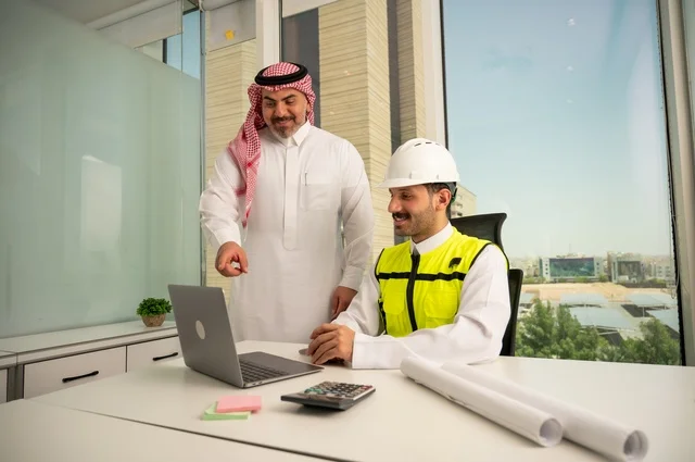 متابعة تنفيذ الخطط الهندسية الموضوعة مسبقا، الشركات المختصة بورشات البناء والمقاولات، التخطيط ووضع اللمسات الأخيرة على المخططات المعمارية، رجل أعمال عربي خليجي سعودي يرتدي الشماغ ومهندس يرتدي الخوذة وسترة الحماية يعملان على مناقشة مخطط الإنشاء وتحديد القياسات بدقة