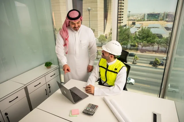 الشركات المختصة بورشات البناء والمقاولات، متابعة تنفيذ الخطط الهندسية الموضوعة مسبقا، التخطيط ووضع اللمسات الأخيرة على المخططات المعمارية، رجل أعمال عربي خليجي سعودي يرتدي الشماغ ومهندس يرتدي الخوذة وسترة الحماية يعملان على مناقشة مخطط الإنشاء وتحديد القياسات بدقة