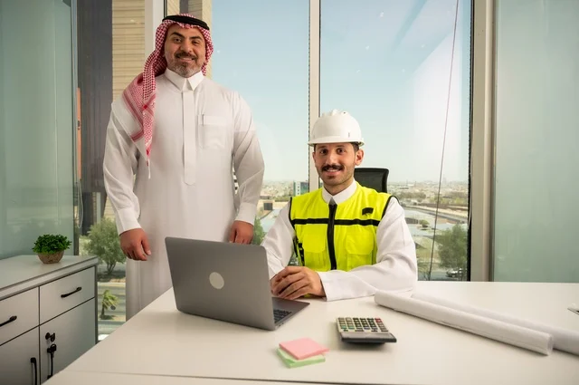 متابعة تنفيذ الخطط الهندسية الموضوعة مسبقا، الشركات المختصة بورشات البناء والمقاولات، التخطيط ووضع اللمسات الأخيرة على المخططات المعمارية، رجل أعمال عربي خليجي سعودي يرتدي الشماغ ومهندس يرتدي الخوذة وسترة الحماية يعملان على مناقشة مخطط الإنشاء وتحديد القياسات بدقة