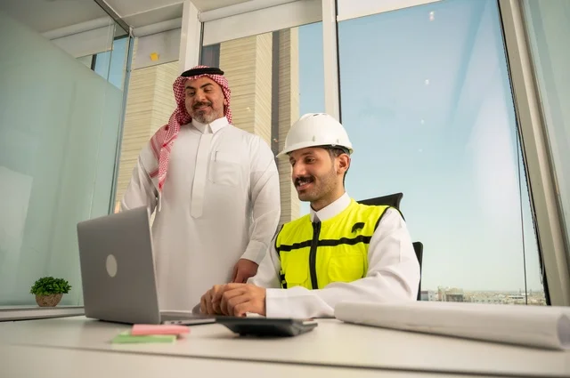 الشركات المختصة بورشات البناء والمقاولات، متابعة تنفيذ الخطط الهندسية الموضوعة مسبقا، التخطيط ووضع اللمسات الأخيرة على المخططات المعمارية، رجل أعمال عربي خليجي سعودي يرتدي الشماغ ومهندس يرتدي الخوذة وسترة الحماية يعملان على مناقشة مخطط الإنشاء وتحديد القياسات بدقة