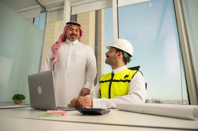 متابعة تنفيذ الخطط الهندسية الموضوعة مسبقا، الشركات المختصة بورشات البناء والمقاولات، التخطيط ووضع اللمسات الأخيرة على المخططات المعمارية، رجل أعمال عربي خليجي سعودي يرتدي الشماغ ومهندس يرتدي الخوذة وسترة الحماية يعملان على مناقشة مخطط الإنشاء وتحديد القياسات بدقة