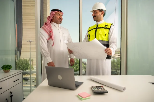 متابعة تنفيذ الخطط الهندسية الموضوعة مسبقا، الشركات المختصة بورشات البناء والمقاولات، التخطيط ووضع اللمسات الأخيرة على المخططات المعمارية، رجل أعمال عربي خليجي سعودي يرتدي الشماغ ومهندس يرتدي الخوذة وسترة الحماية يعملان على مناقشة مخطط الإنشاء وتحديد القياسات بدقة