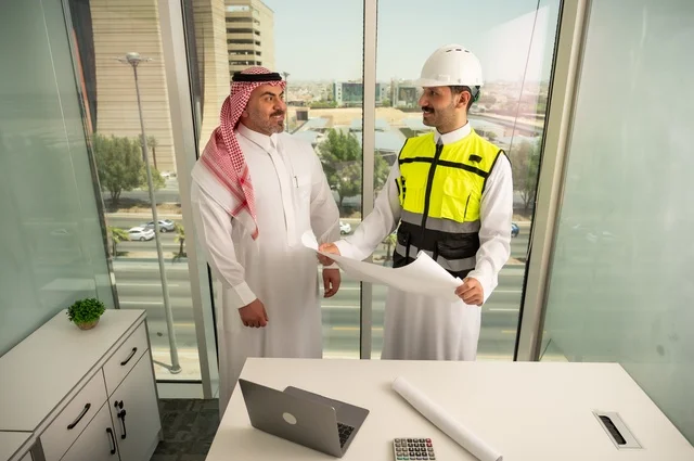 متابعة تنفيذ الخطط الهندسية الموضوعة مسبقا، الشركات المختصة بورشات البناء والمقاولات، التخطيط ووضع اللمسات الأخيرة على المخططات المعمارية، رجل أعمال عربي خليجي سعودي يرتدي الشماغ ومهندس يرتدي الخوذة وسترة الحماية يعملان على مناقشة مخطط الإنشاء وتحديد القياسات بدقة