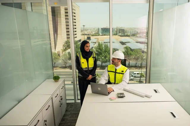 دمج التكنلوجيا مع العمل، التعاون لإنجاز العمل بسرعة ودقة، التدقيق والإشراف على المشاريع الهندسية من مكتب هندسي, مهندسان عربيان خليجيان سعوديان يرتديان خوذة الحماية وسترة العمل و يقومان بدراسة و تقييم الخطط الهندسية من خلال الحاسوب المحمول